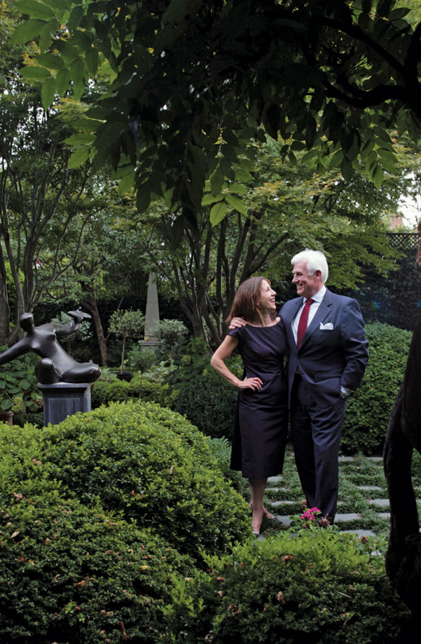 Irene and John Danilovich in their garden, where a Haddonstone obelisk is centered at the rear wall between a pergola-like gardening shed and a lap pool. 