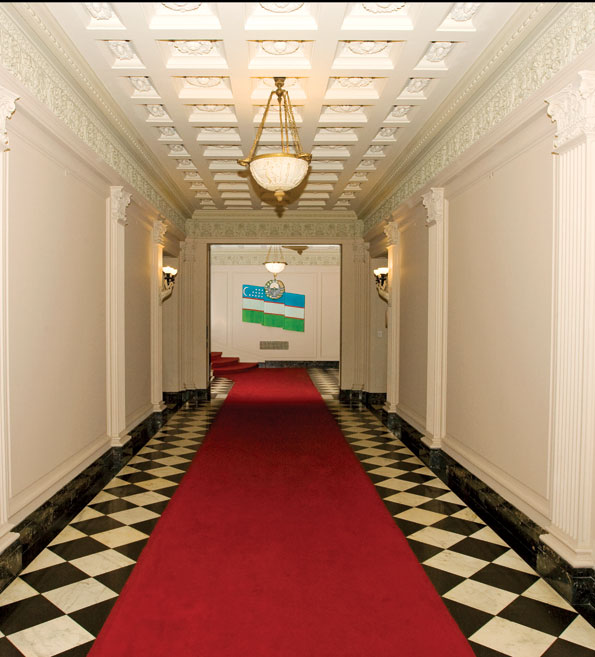  The expansive Entry Hall, which features fluted pilasters, an elaborate coffered ceiling, ornate carvings, and a checkerboard marble floor, connects to the grand staircase in the rear. The bas-relief flag of the Republic of Uzbekistan dominates the background